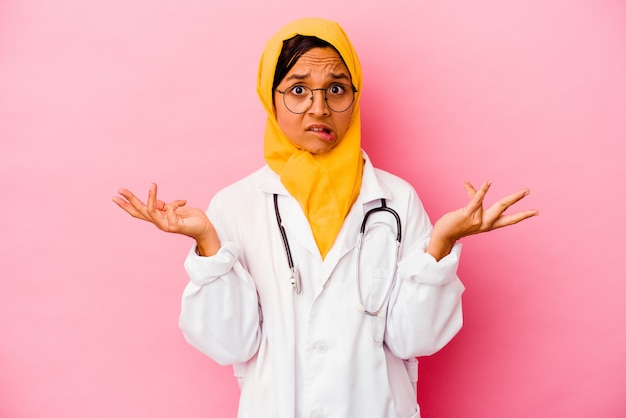 Jonge artsen moslimvrouw geïsoleerd op roze achtergrond verward en twijfelachtig schouders ophalen om een kopie ruimte vast te houden.