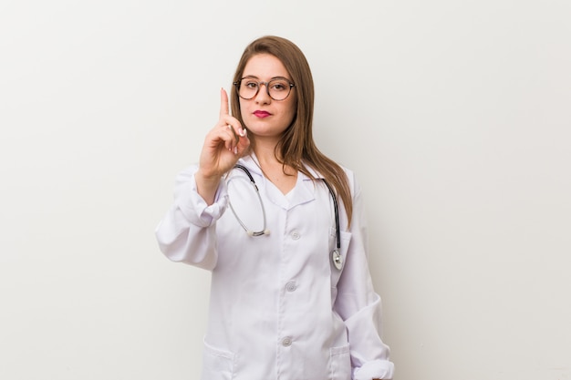 Jonge artsenvrouw tegen een witte muur die nummer één met vinger toont.