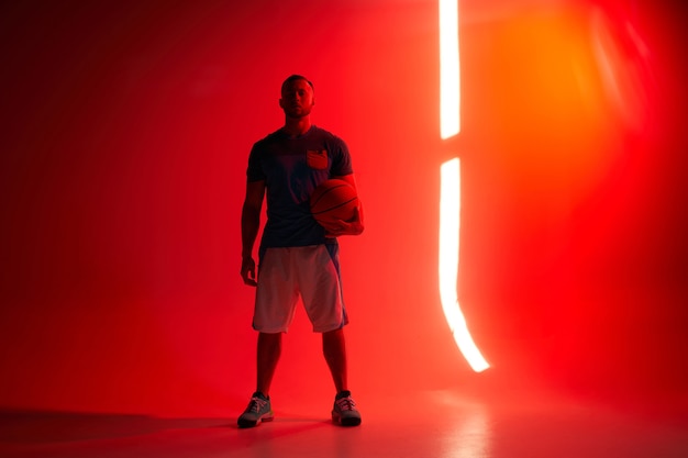 Jonge atletische basketbalspeler staat met de bal in de ene hand met achtergrondverlichting