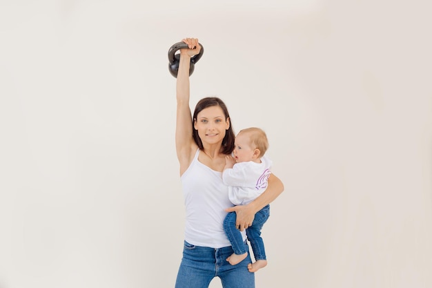 Jonge atletische glimlachende vrouw die de kettlebell optilt en de kracht van een kleine baby op een witte achtergrond vasthoudt