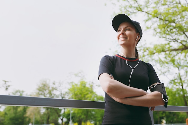 Jonge atletische lachende vrouw in zwart uniform, pet met koptelefoon luisteren naar muziek, staande handen gevouwen houden voor of na het hardlopen, trainen in stadspark buitenshuis