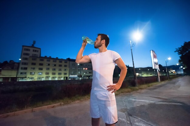 jonge atletische man die water drinkt na een nachtelijke hardloopsessie in de stad