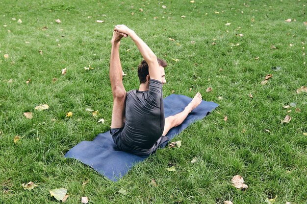 Jonge atletische man in sportkleding die yoga doet in het park. Oefen asana buitenshuis. Mensen die zich uitstrekken op groen gras met yogamat. Fitness en gezonde levensstijl. Concept van meditatie