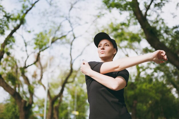 Jonge atletische mooie brunette vrouw in zwart uniform, pet doet sport rekoefeningen, warming-up voor hardlopen of training, staande in stadspark buitenshuis