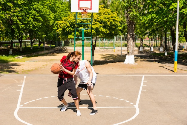 Jonge atletische paar spelen basketbal samen op buitenbaan in weelderig groen Park