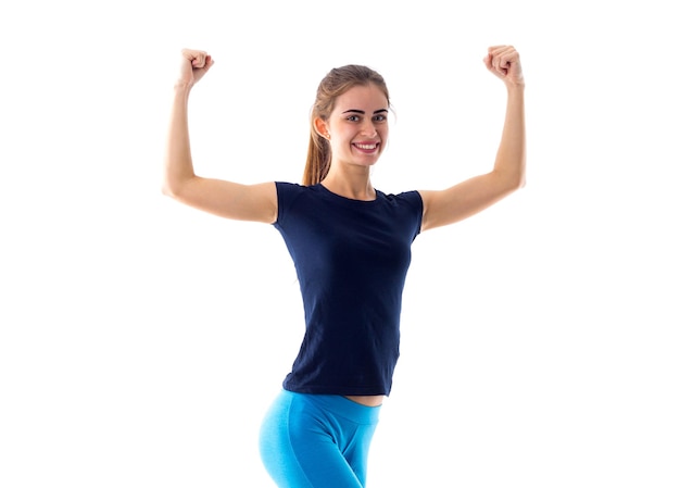 Jonge atletische vrouw in blauw T-shirt en beenkappen die biceps op witte achtergrond in studio tonen