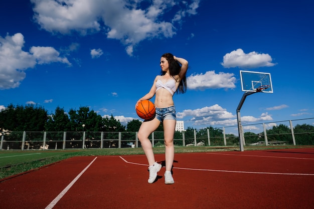 Jonge atletische vrouw, in top en joggingbroek, spelen met bal op basketbalveld buitenshuis.