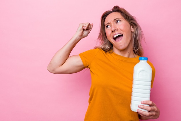 Jonge australische vrouw die een fles melk houdt die op roze achtergrond wordt geïsoleerd en vuist opheft na een overwinning, winnaarconcept.