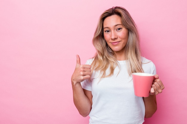 Jonge australische vrouw die een roze mok houdt die op roze achtergrond wordt geïsoleerd die en duim glimlacht opheft