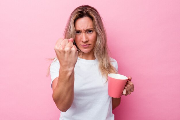 Jonge Australische vrouw die een roze mok houdt die op roze achtergrond wordt geïsoleerd die vuist toont aan camera, agressieve gezichtsuitdrukking.