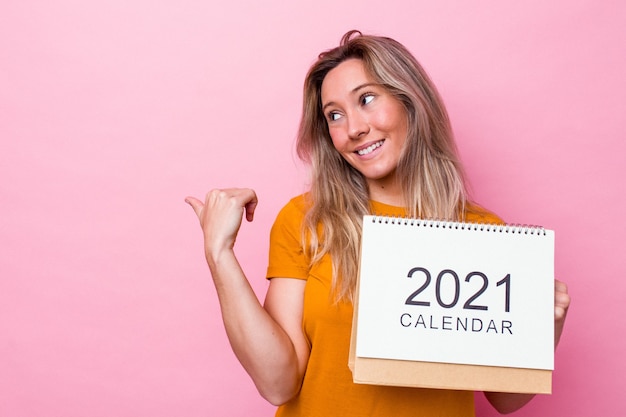 Jonge australische vrouw met een kalender geïsoleerd op roze achtergrond wijst met duimvinger weg, lachend en zorgeloos.