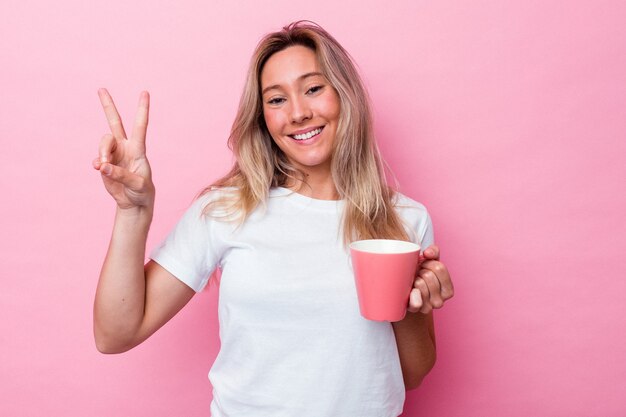 Jonge australische vrouw met een roze mok geïsoleerd op roze achtergrond vrolijk en zorgeloos met een vredessymbool met vingers.