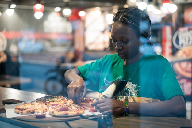 Jonge authentieke Afro-Amerikaanse vrouw die pizza eet in het restaurant van de nachtstad