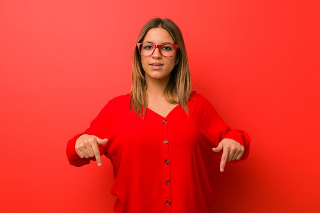 Jonge authentieke charismatische echte mensenvrouw tegen een muur wijst naar beneden met vingers, positief gevoel.