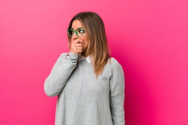 Jonge authentieke charismatische echte mensenvrouw tegen een nadenkende muur kijkend aan een exemplaarruimte die mond behandelen met hand.