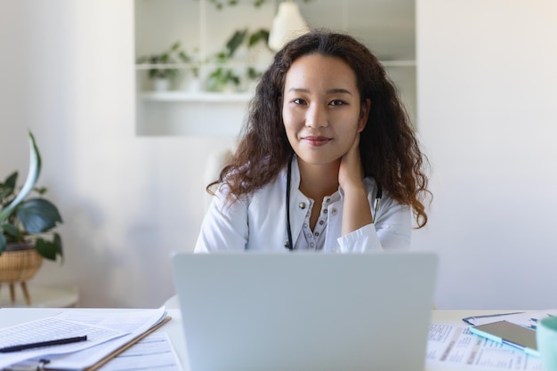 Jonge Aziatische arts die laptop gebruikt en aan een bureau zit Vrouw professionele arts met witte jas en stethoscoop die op de computer op de werkplek werkt