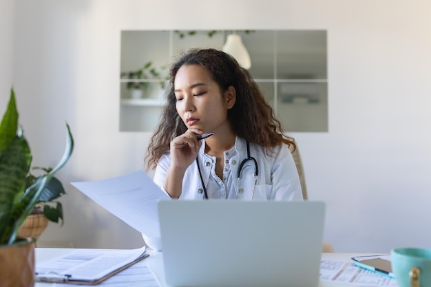 Jonge Aziatische arts die laptop gebruikt en aan een bureau zit Vrouw professionele arts met witte jas en stethoscoop die op de computer op de werkplek werkt