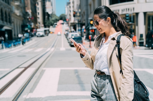 Jonge Aziatische Chinese zakenvrouw op zonnige drukke stadsstraat in San Francisco Amerika. vrouw manager sms't op mobiele telefoon lopen op de weg in de stad. lachende kantoordame in zonnebril gebruikt mobiele telefoon