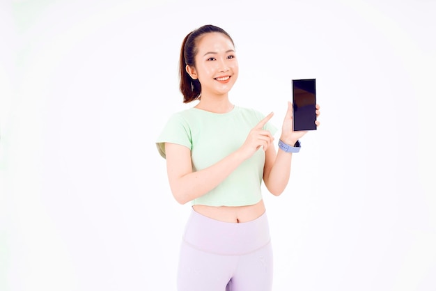 Jonge Aziatische dame toont leeg smartphonescherm met positieve uitdrukking glimlacht breed gekleed in casual kleding, voelt zich gelukkig op blauwe achtergrond Mobiele telefoon met wit scherm in vrouwelijke hand