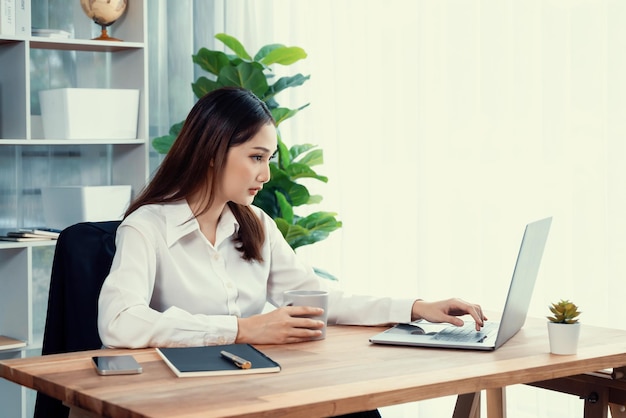 Jonge Aziatische enthousiaste zakenvrouw aan een modern kantoor bureau met een laptop om te werken met een kop koffie Diligente en aantrekkelijke kantoorvrouw die aan een computer notebook werkt in haar kantoorwerkruimte