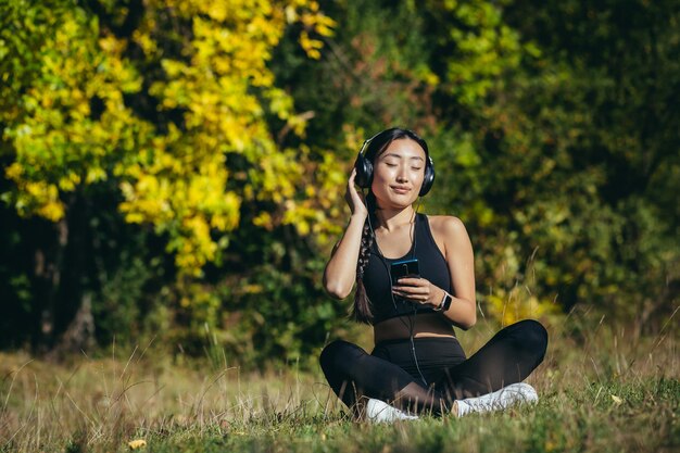 Jonge Aziatische fit vrouw zittend op mat in lotuspositie mediteren ontspannen en luisteren naar muziek buitenshuis. Happy Girl-vrouw geniet van de natuur met een koptelefoon in het bos of park. Genezing met geluiden