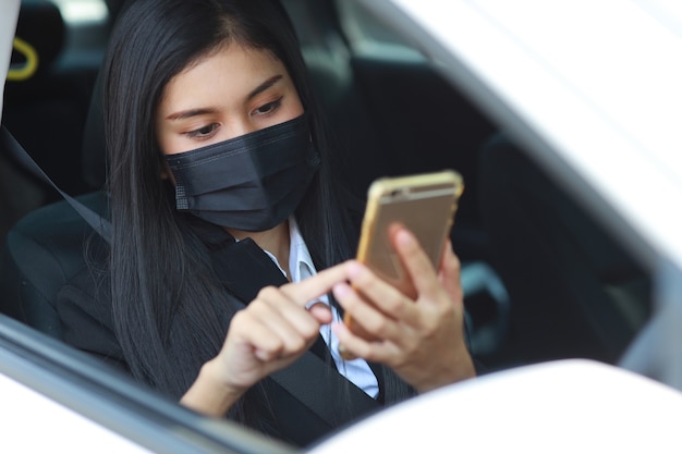 Jonge aziatische gezonde vrouw in zwart zakelijk pak met beschermend masker voor gezondheidszorg in de auto en het gebruik van smartphone en autorijden. Nieuw normaal en social distancing concept