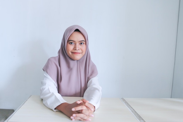 Jonge Aziatische islamvrouw met hoofddoek zit op tafel met hand in hand en glimlacht islam Indonesische vrouw op grijze achtergrond