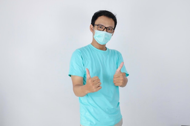 Jonge Aziatische man die een medisch masker draagt, geeft een goed handteken met zelfverzekerde gebaren Indonesische man met een blauw shirt