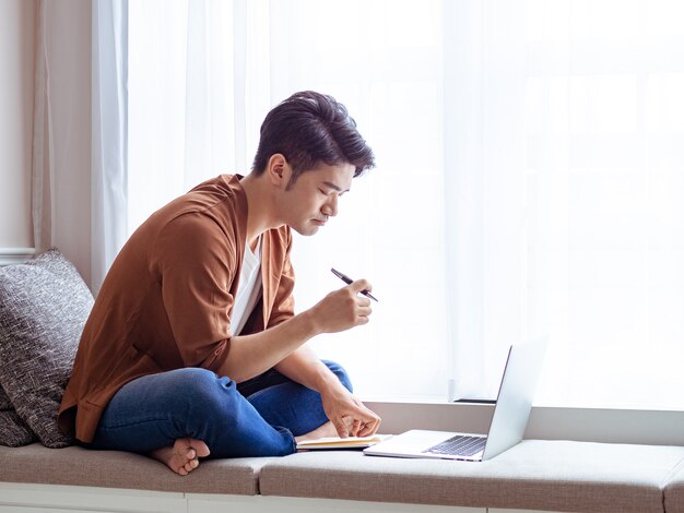 Jonge aziatische man die in notitieboekje schrijft en laptop gebruikt.