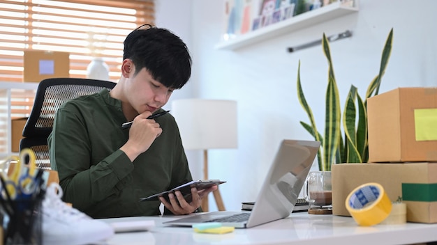 Jonge Aziatische man die online bestelling op digitale tablet controleert en pakketdozen voorbereidt voor levering aan klanten