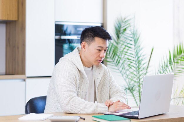 Jonge aziatische man die thuis werkt op een laptopcomputer die aan een tafel in de keuken zit