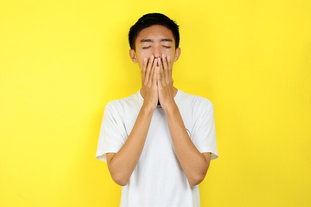 Jonge aziatische man die verdrietig is, sluit zijn gezicht met zijn hand, geïsoleerd op gele achtergrond