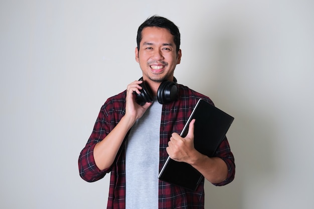 Jonge Aziatische man die zelfverzekerd glimlacht terwijl hij een hoofdtelefoon in zijn nek draagt en een laptop vasthoudt