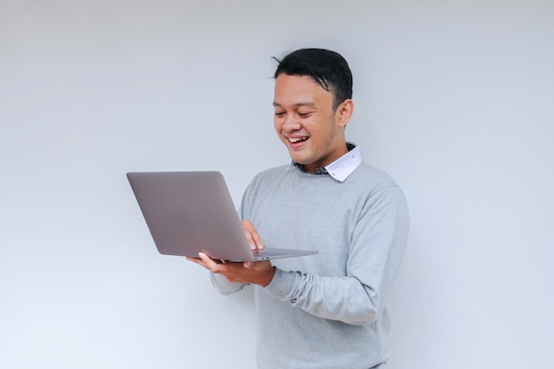 Jonge Aziatische man die zich gelukkig voelt en glimlacht als hij staat en op een laptop werkt Indonesische man met een grijs shirt