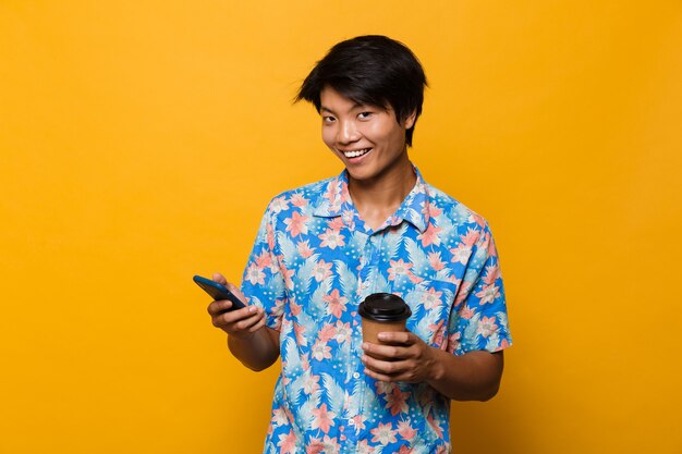 Jonge Aziatische man geïsoleerd over gele ruimte met behulp van mobiele telefoon koffie drinken.