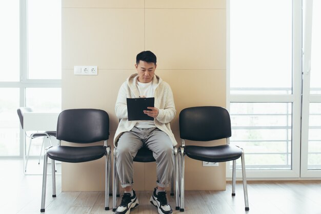 Jonge aziatische man in wachtkamer voor interview of vergadering met papier terwijl hij aan de stoel zit, vult een cv-vragenlijst in op kantoor
