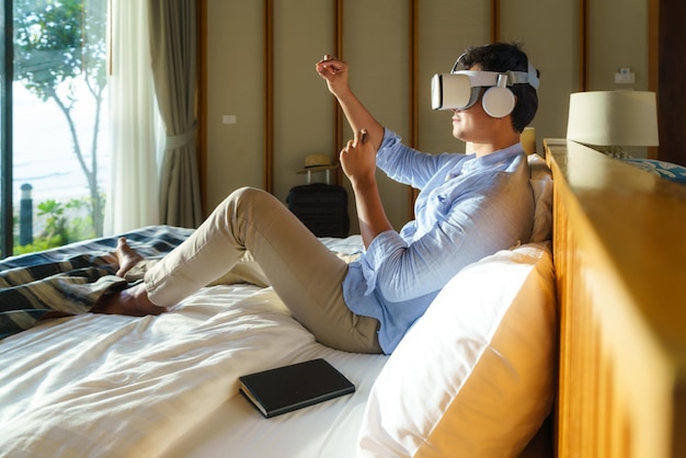 Jonge Aziatische man liggend op bed en met behulp van virtual reality-bril voor zakelijke bijeenkomst tijdens zijn reizen zomervakantie vakantie.