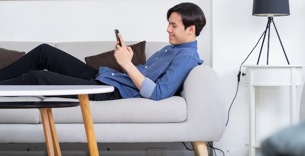 Jonge Aziatische man liggend op de bank en met behulp van telefoon