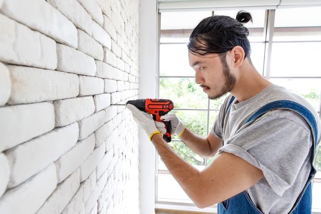 Jonge Aziatische man met behulp van elektrische boor op witte bakstenen muur in de kamer, concept voor huisverbetering.