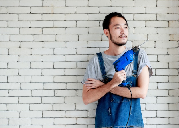 Jonge Aziatische man met een boormachine die voor een witte bakstenen muur staat, glimlacht en naar buiten kijkt, concept voor thuis DIY