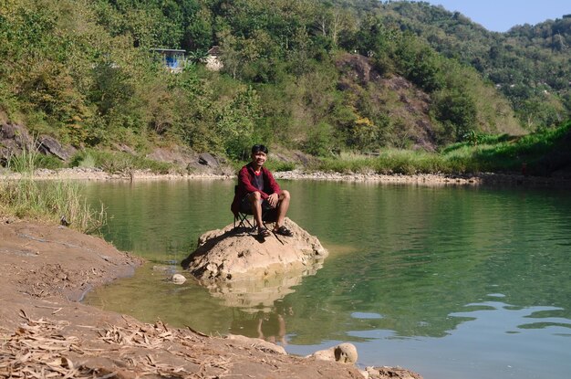 Jonge Aziatische man met een rood jasje zit ontspannen bij de rivier.