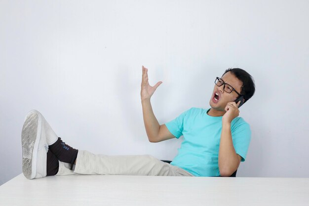 Jonge Aziatische man met geschokt gezicht op mobiele telefoon met been op tafel Indonesische man met blauw shirt