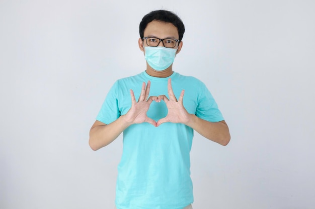 Jonge Aziatische man met masker voor bescherming tegen ziekte met liefde Korea bord met geïsoleerde witte achtergrond Corona virus concept Indonesische man met blauw shirt