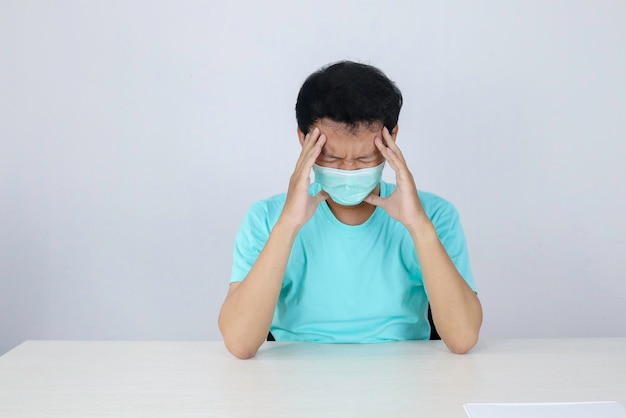 Jonge aziatische man met medisch masker lijdt aan ernstige hoofdpijn die vingers drukt tegen slapende ogen om pijn te verlichten met hulpeloze gezichtsuitdrukking