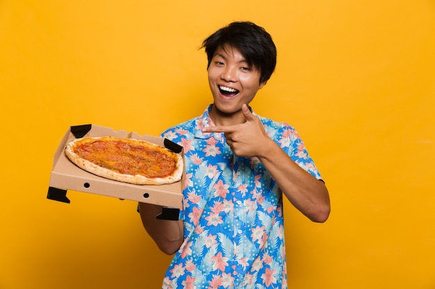Foto jonge aziatische man staande geïsoleerd over gele ruimte met pizza wijzen.