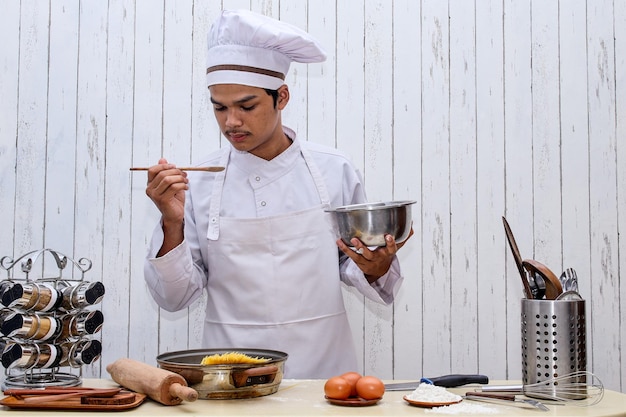 Jonge Aziatische mannelijke chef-kok in witte toque en schort met kom en houten spatel die het eten proeft