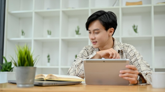 Jonge Aziatische mannelijke freelancer die digitale tablet-touchpad gebruikt tijdens het lezen van een boek