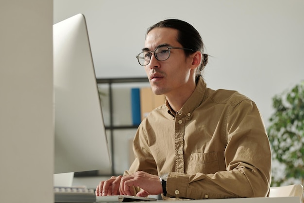 Jonge Aziatische mannelijke ondernemer kijkend naar het computerscherm op de werkplek