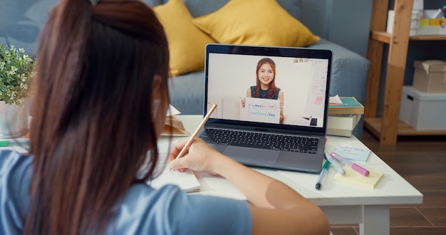 Foto jonge aziatische meid met casual gebruik computer laptop videogesprek online leren met leraar schrijf lezing notebook woonkamer thuis