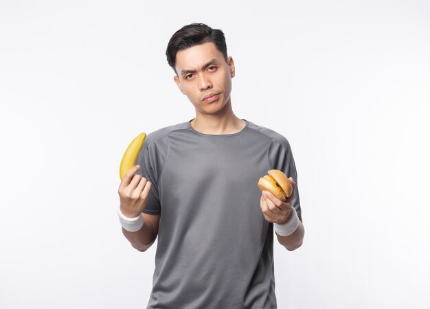 Jonge Aziatische mens die in sportuitrustingen banaan en hamburger met geïsoleerd twijfelsgezicht houden